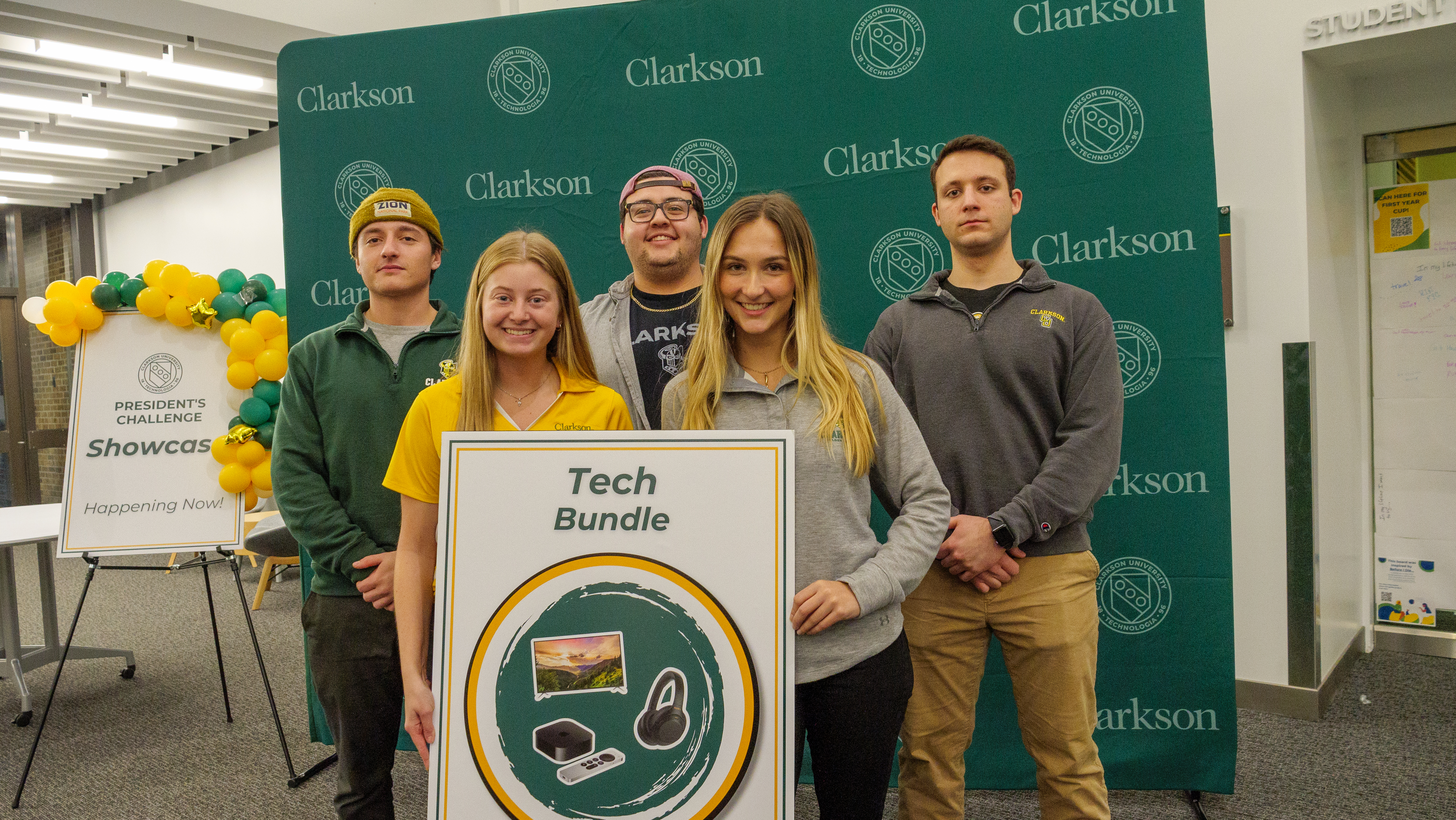Five Clarkson students pose in front of a branded banner with a poster showcasing their prize for winning the President’s Challenge. 