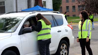 Two Clarkson University occupational therapy students in fluorescent green vests approach a woman in a small white SUV wagon.