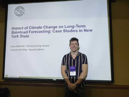 Devin Hodoroski, in striped polo shirt, posing in front of a PowerPoint slide on a screen.