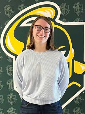 Waist-up portrait of Lindsey Penet wearing white shirt and standing in front of the Golden Knights athletic logo.