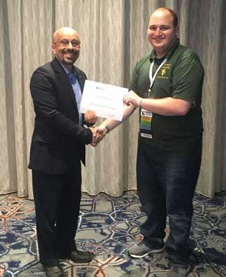 A man with glasses in a suit handing a paper certificate to Michael Buchwald, wearing a green polo shirt.