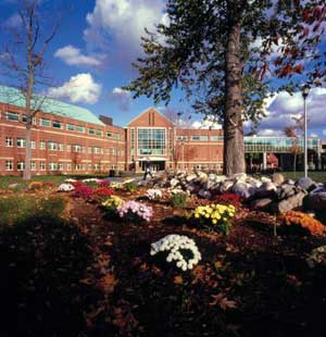 Exterior of Bertrand H. Snell Hall on Clarkson University's hill campus with flowers and a tree in the foreground and a partly cloudy blue sky.
