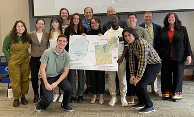 Group of 14 people posing with a mounted map of the Adirondack Park