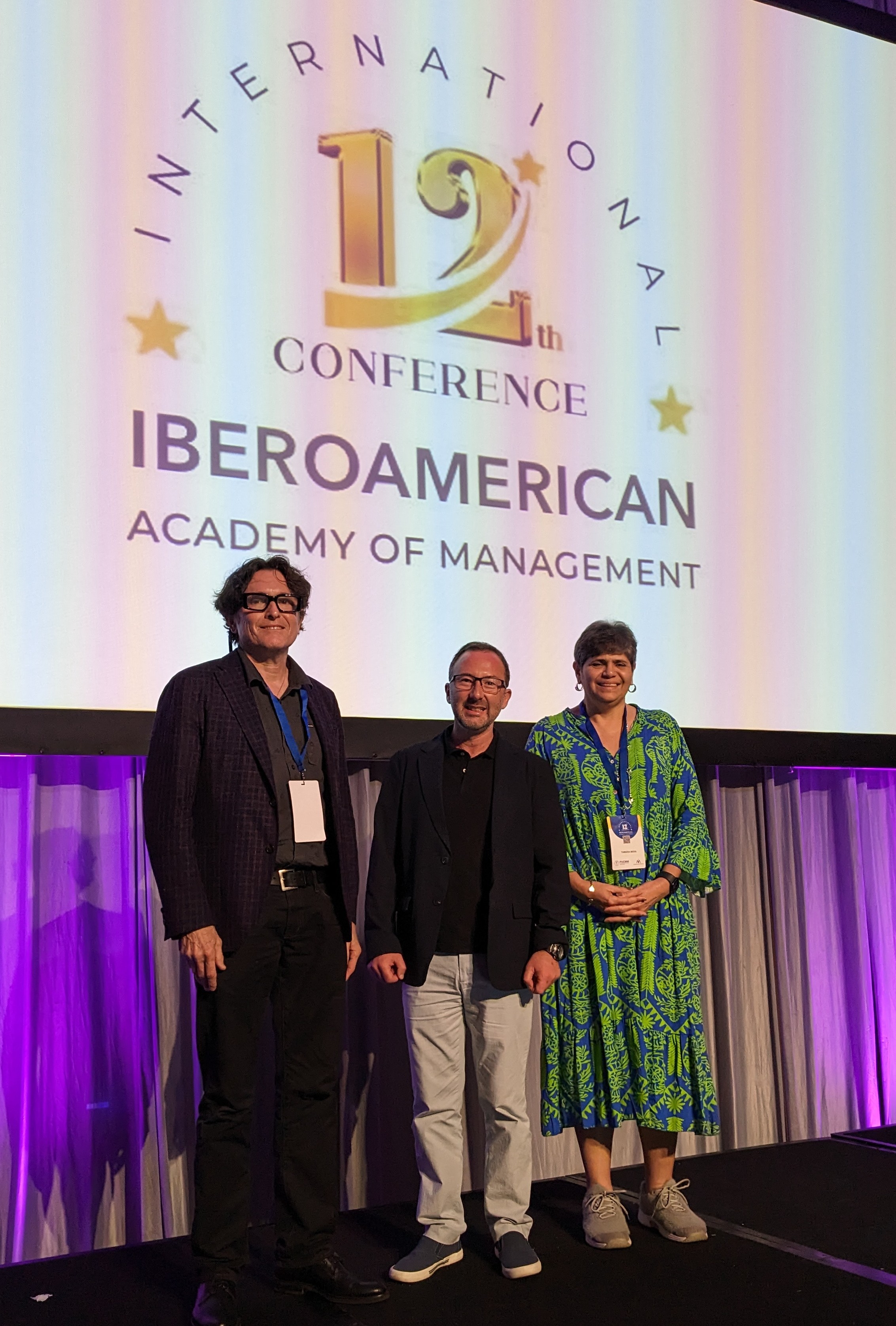 Christian Felzensztein stands between two presenters on stage in front of a screen with the logo for the 12th conference of the International Iberoamerican Academy of Management. 