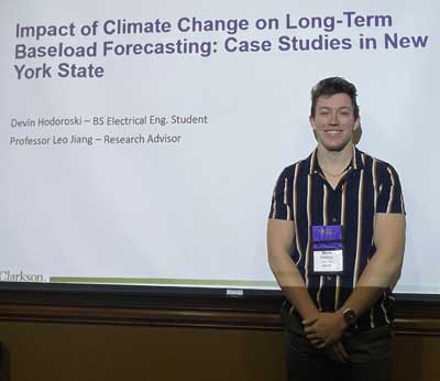 Waist-up portrait of Devin Hodoroski in a vertically striped shirt standing in front of a PoerPoint screen reading "Impact of Climate Change on Long-Term Baseload Forecasting: Case Studies in New York State"
