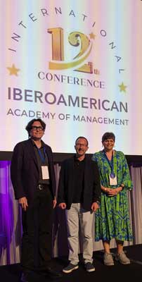 Christian Felzensztein stands between two presenters on stage in front of a screen with the logo for the 12th conference of the International Iberoamerican Academy of Management.
