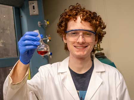 Matthias Schmidt wearing a white lab coat, safety goggles and a blue glove, holding a beaker with a red liquid