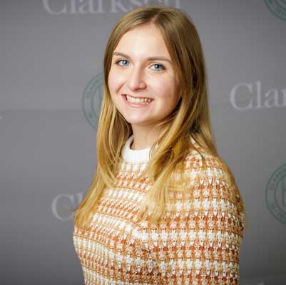 Chest-up portrait of Nicolena Fazio in a striped, tan sweater, and a Clarkson logo background