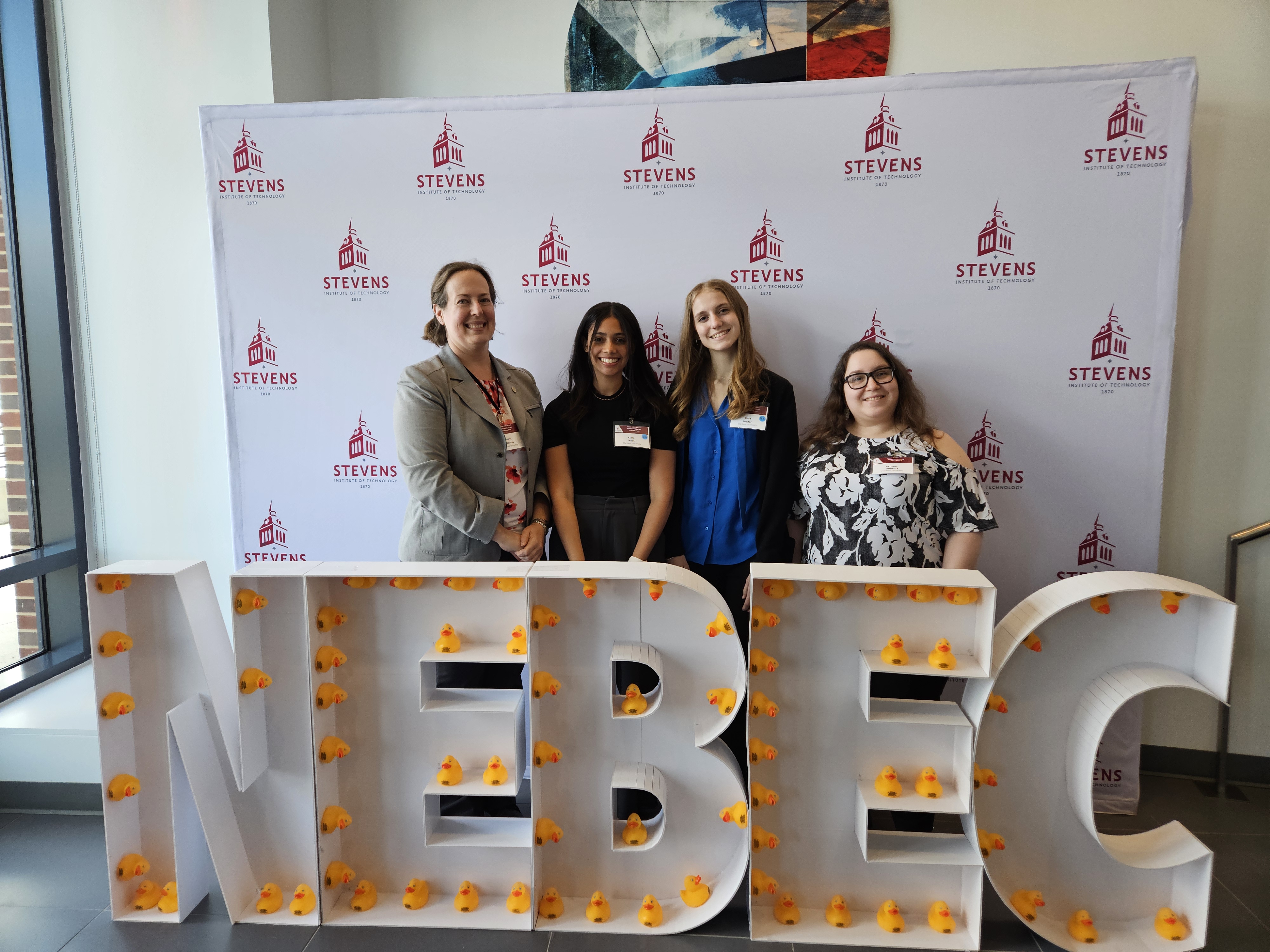 Two students and two professors stand in front of a Stevens Institute backdrop with large cardboard letters spelling NEBEC in the foreground. 