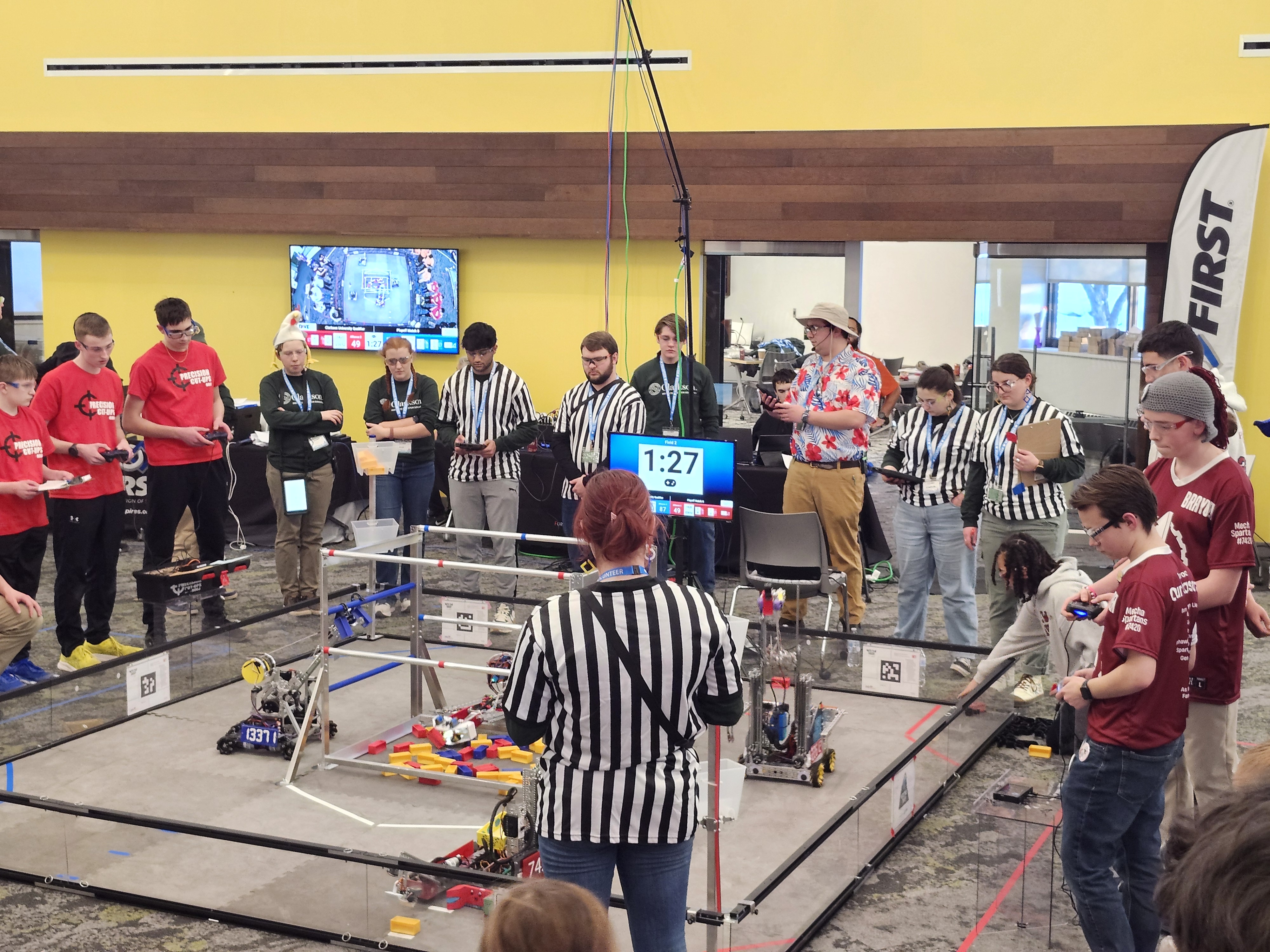 Several students in team t-shirts and referees gather around a robotics ring.