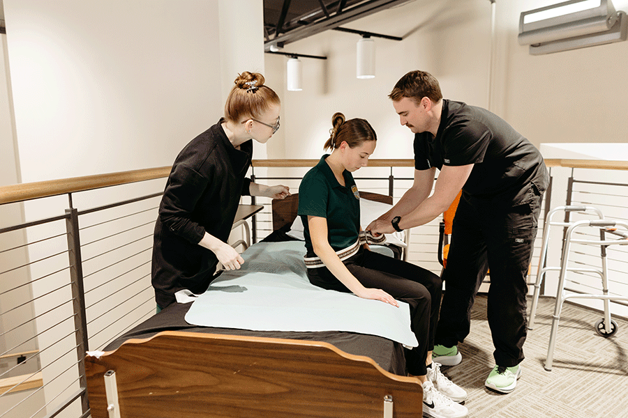 Two health science studies practice on a third student serving as a patient with an occupational therapy belt on a twin size bed in Clarkson University's Sim Lab.