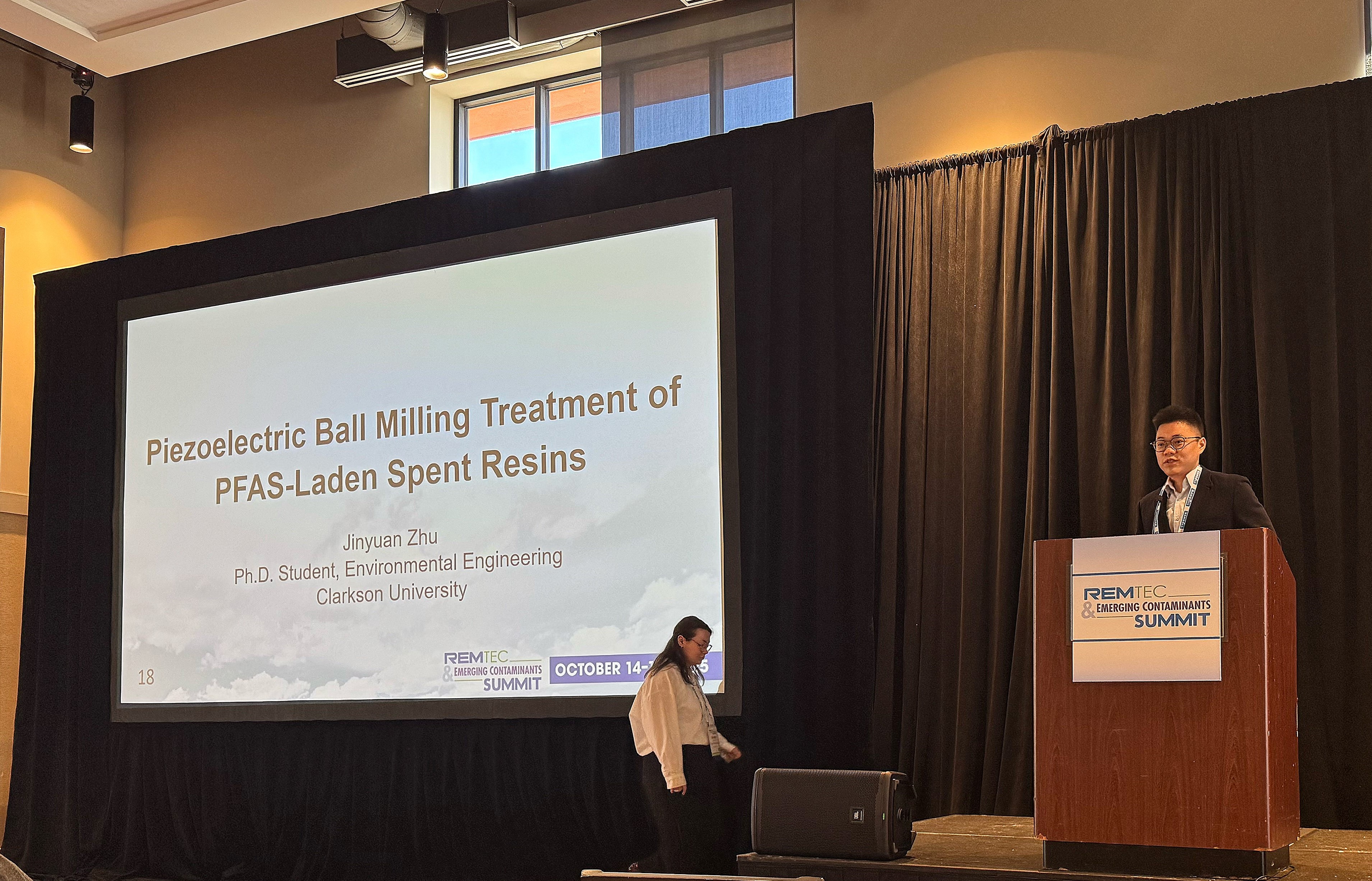 A student stands at a podium on stage presenting research displayed on a large projector screen next to him.