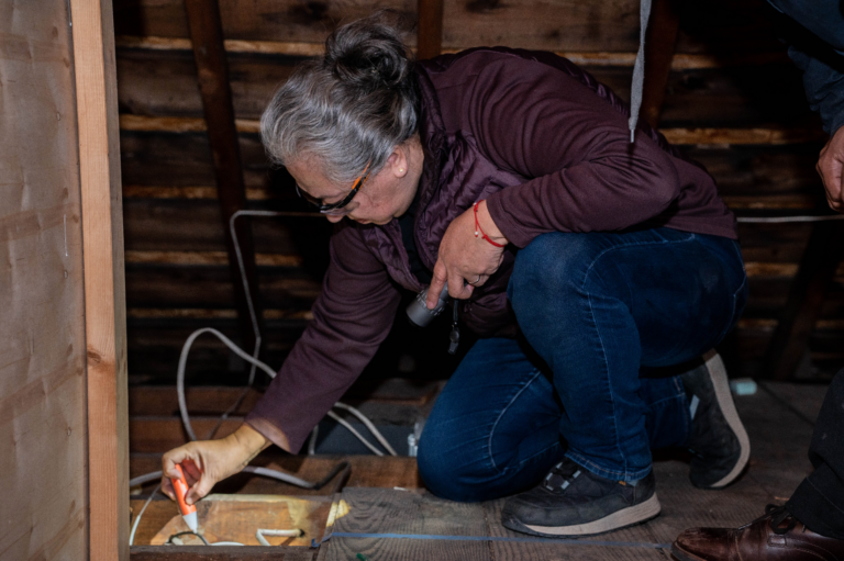 An older woman kneels down and uses an electric current tester on a wire in an attic space