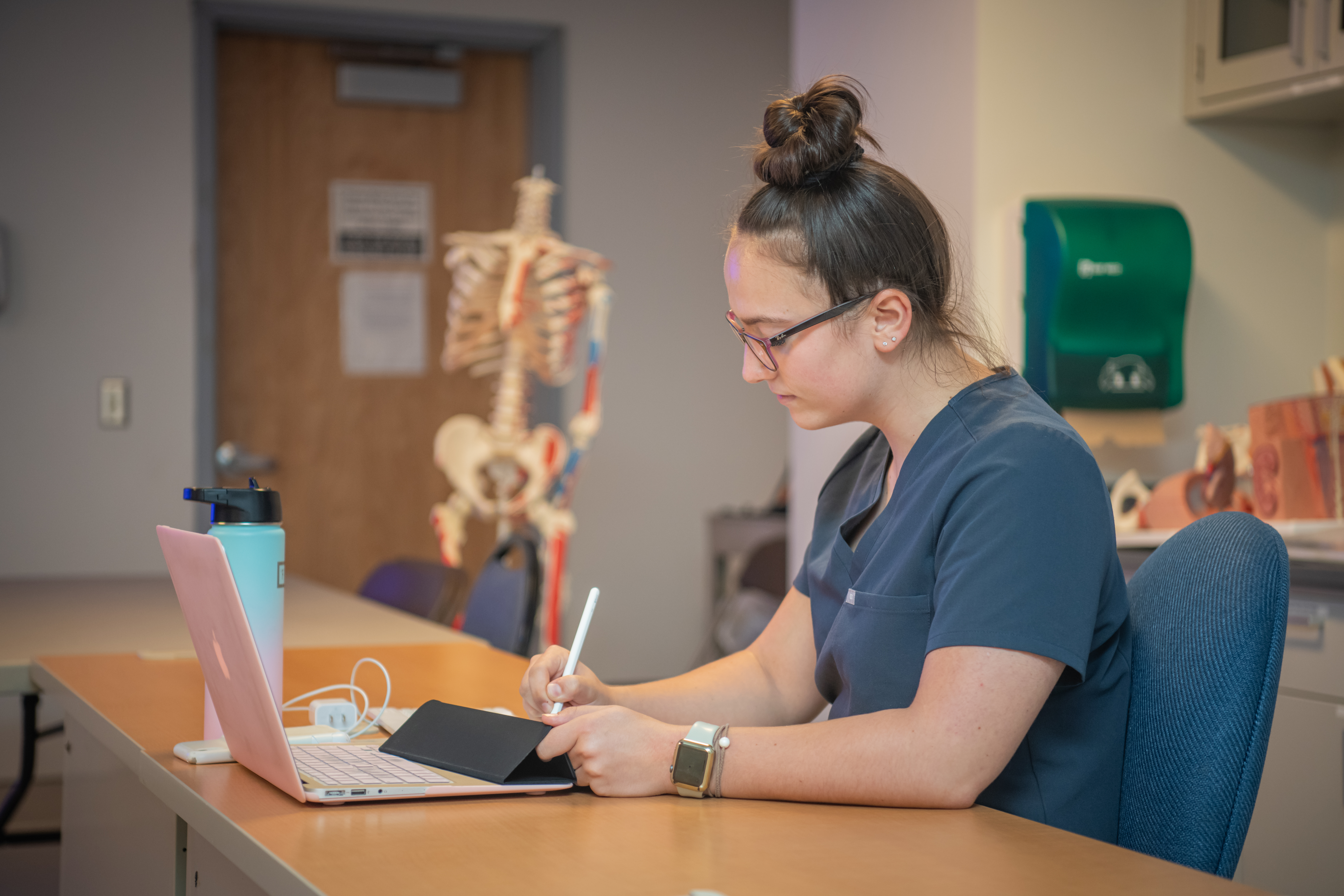 A student writes on a tablet with a laptop in front of her, in a medical office setting.