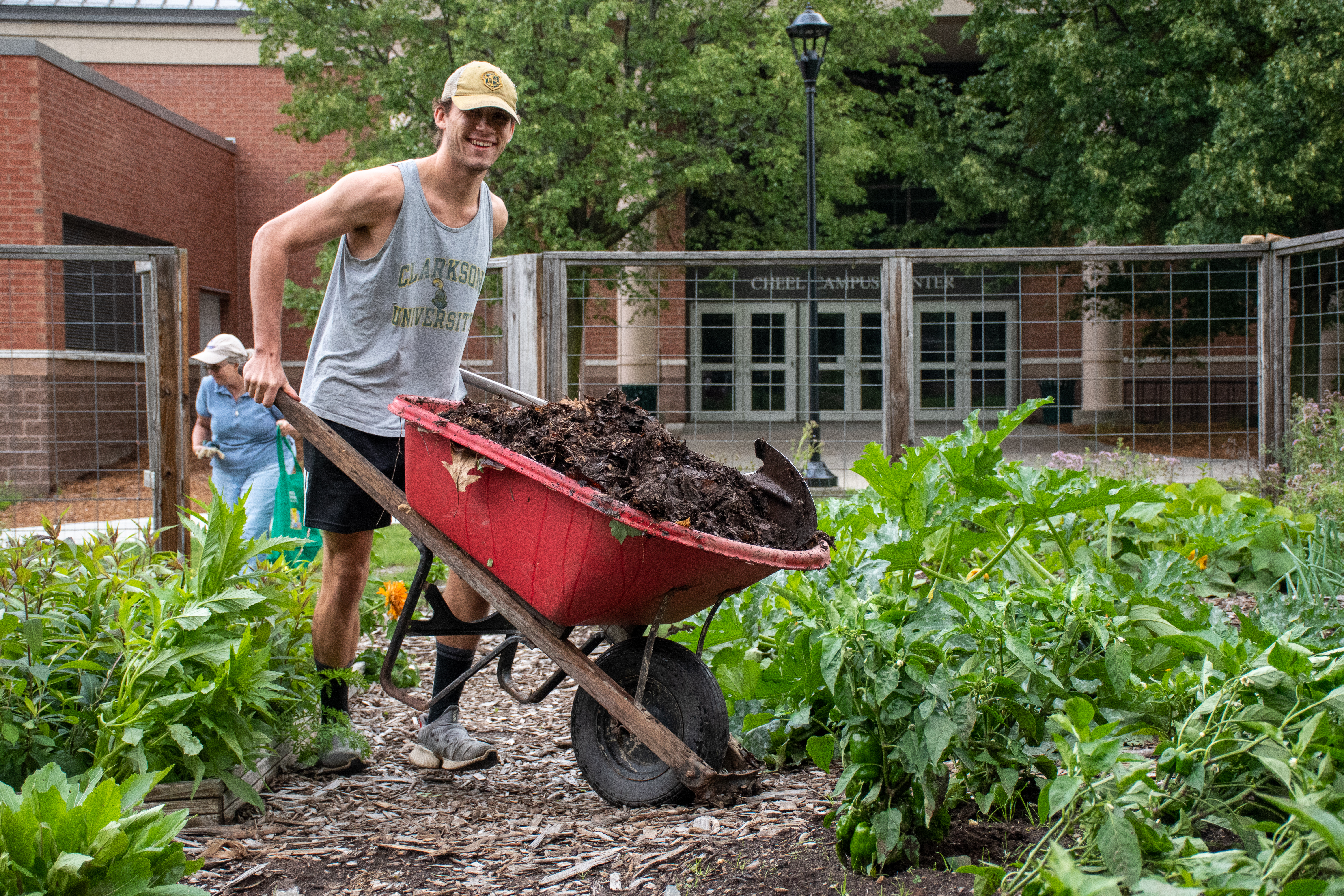 A young man smiles for the camera as he pushes a wheel barrow through a garden