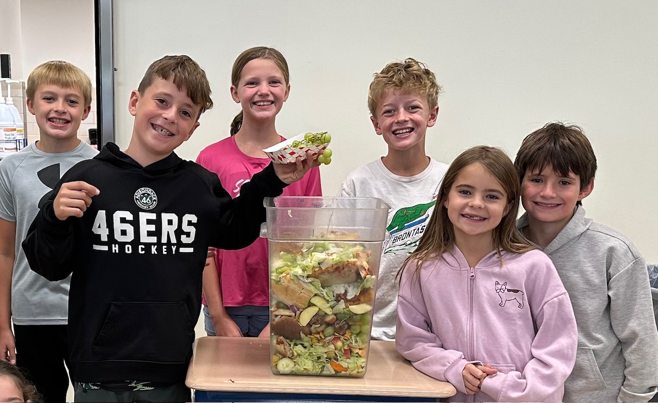 Several elementary school students pose for a photo surrounding a large, clear compost bucket, with one child ready to dump compostable items into the bucket. 