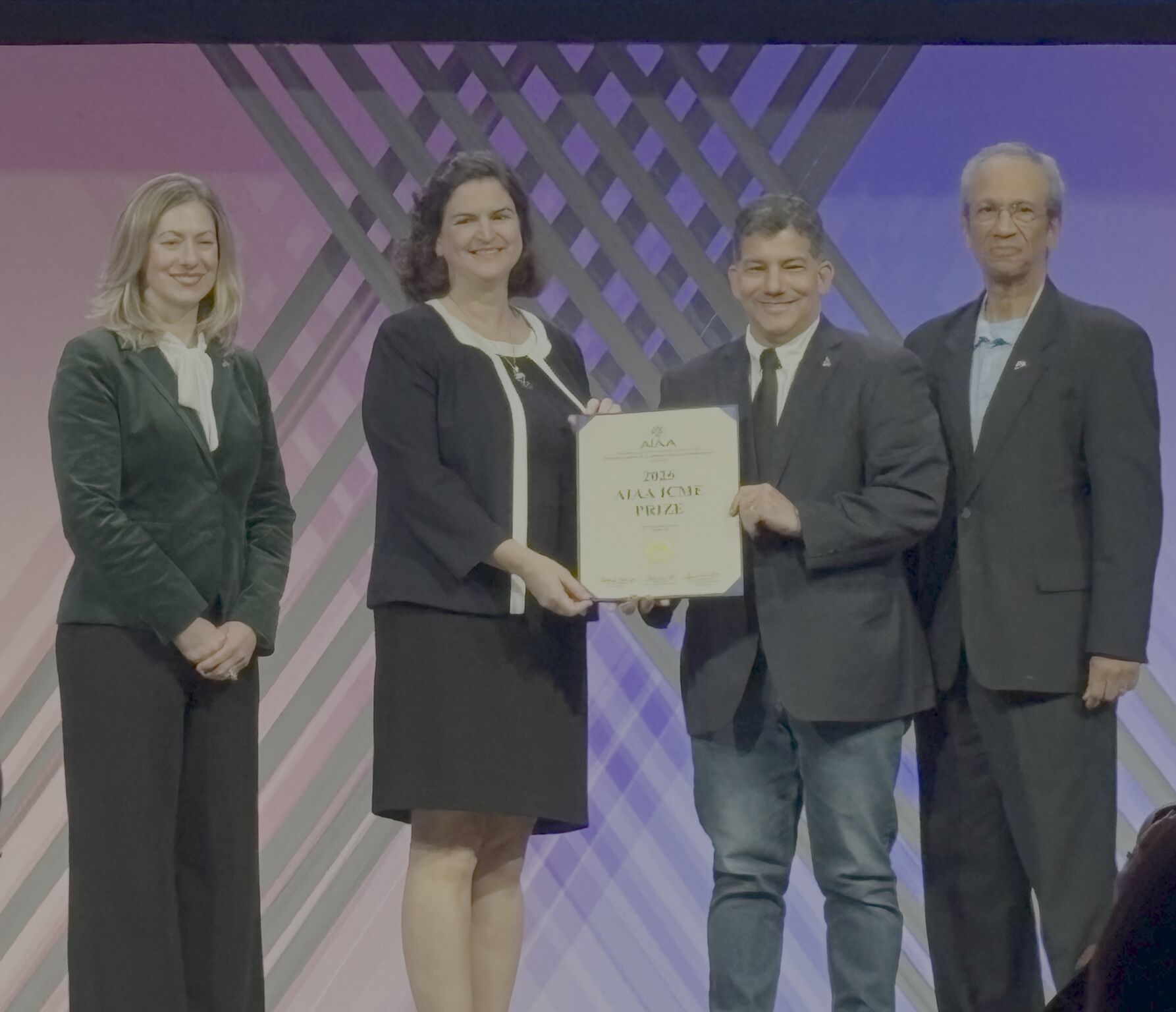 four people pose on a conference stage, while the two in the middle grip a paper award