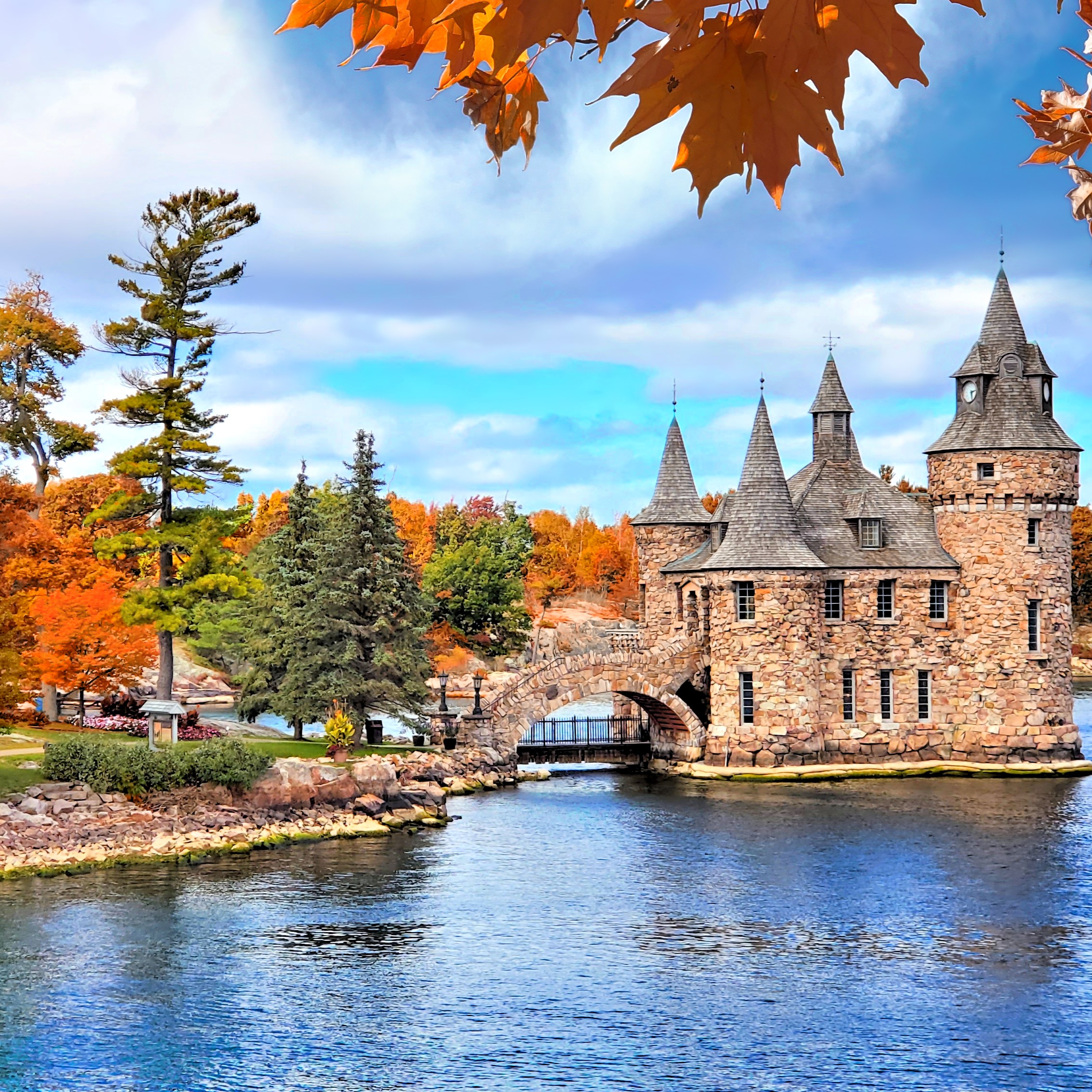The historic stone Power House at Boldt Castle on Heart Island, surrounded by vibrant orange and yellow autumn foliage and blue water under a partly cloudy sky.