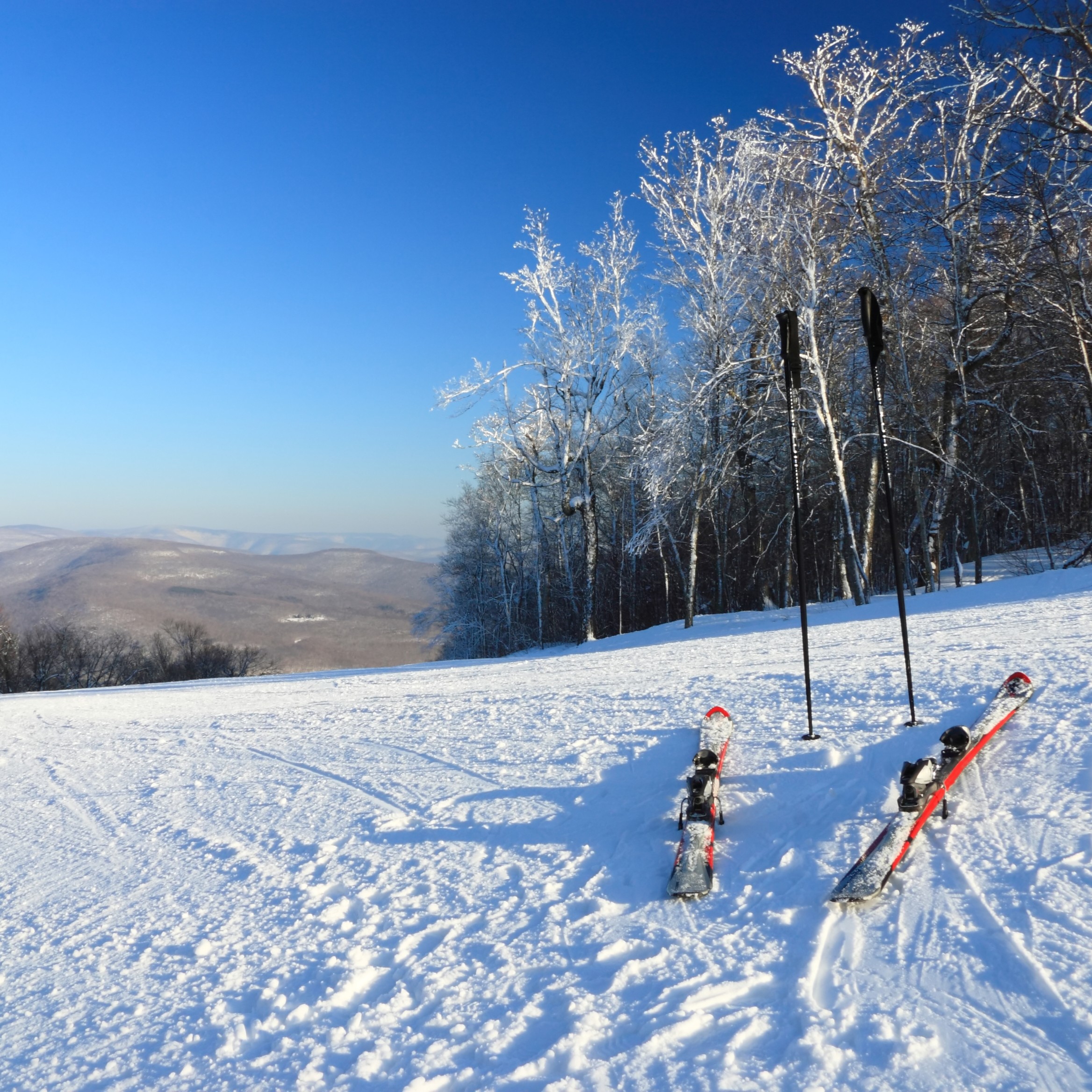 A pair of red and black downhill skis and black ski poles resting on a groomed, snow-covered slope at a ski resort under a clear blue sky, overlooking a valley of rolling hills