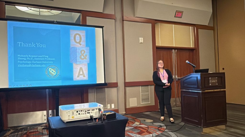 A student presents from a podium in a conference center next to a projector displaying the final thank you slide of her research presentation. 