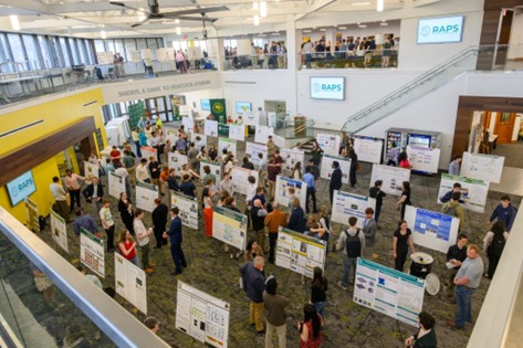A group of students, faculty, and staff stand around poster presentations in an auditorium-sized space.