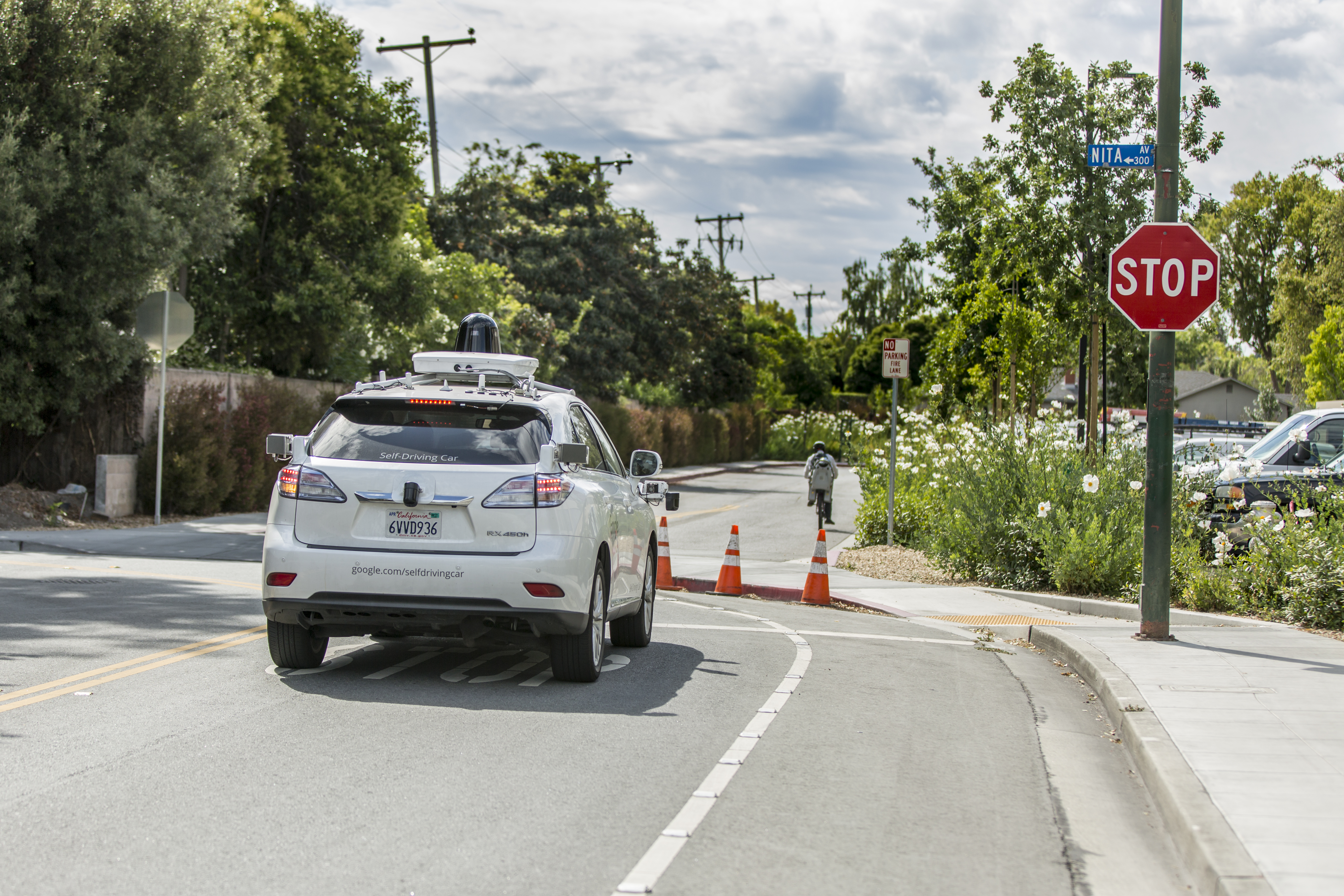 A white self-driving car equipped with rooftop sensors and cameras drives along a suburban street toward a stop sign. The road is lined with trees, utility poles, parked cars, and flowering plants, with orange traffic cones marking the bike lane near the intersection.