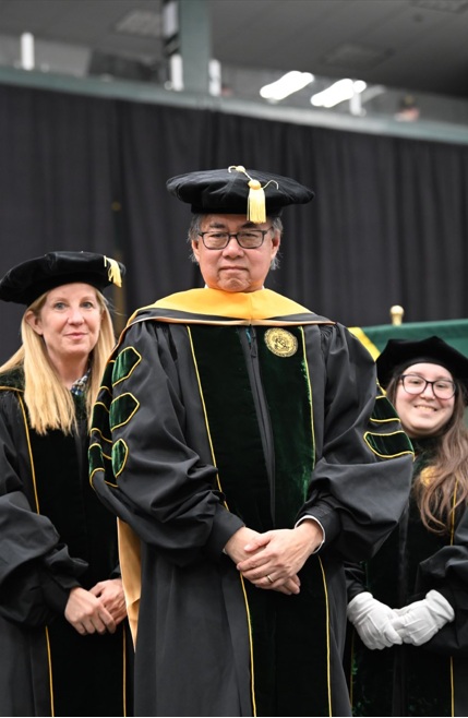 a man stands posed for a photo after being hooded in his commencement regalia on stage.