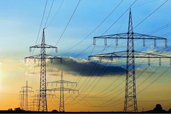 Several tall electrical transmission towers carrying high-voltage power lines stretch across the landscape at sunset, silhouetted against a sky with blue and orange tones and scattered clouds.