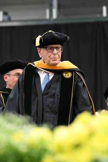 An elderly man crosses a stage in commencement regalia