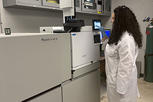 A researcher in a white lab coat and blue gloves operates a touchscreen on a large gas chromatography mass spectrometer instrument in a university laboratory.