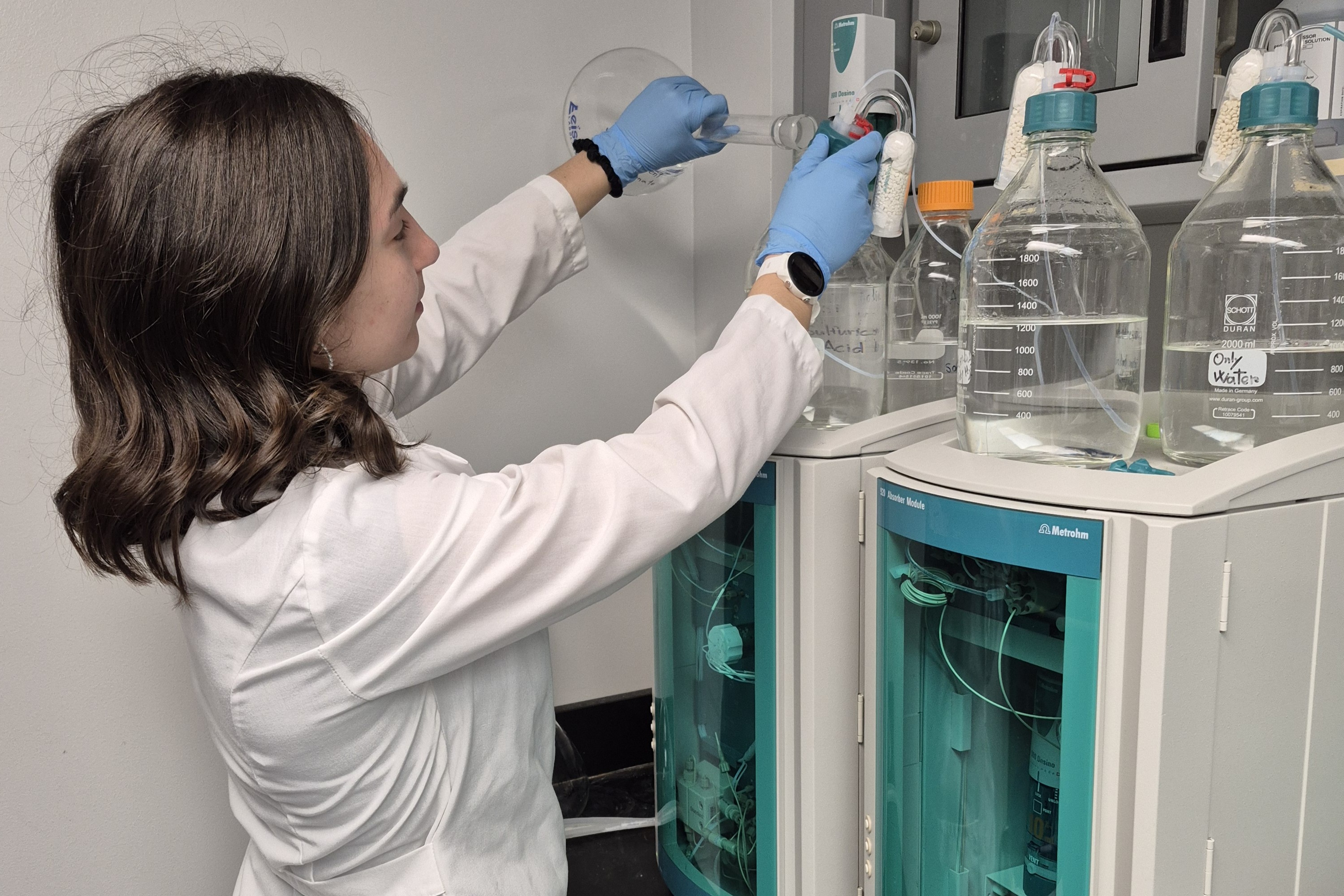 A female Clarkson University researcher wearing a white lab coat and blue gloves uses a pipette to prepare a solution into a glass bottle. In the foreground, stacked analytical instruments with teal accents and glass bottles containing clear liquids sit on a laboratory counter.