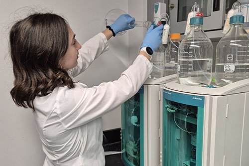 A researcher wearing a white lab coat and blue gloves carefully adjusts glass solvent bottles on top of an ion chromatography system in a university laboratory.