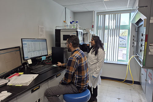 Two researchers collaborating in a laboratory, with one sitting at a computer workstation and another standing in a white lab coat reviewing data on the monitor.