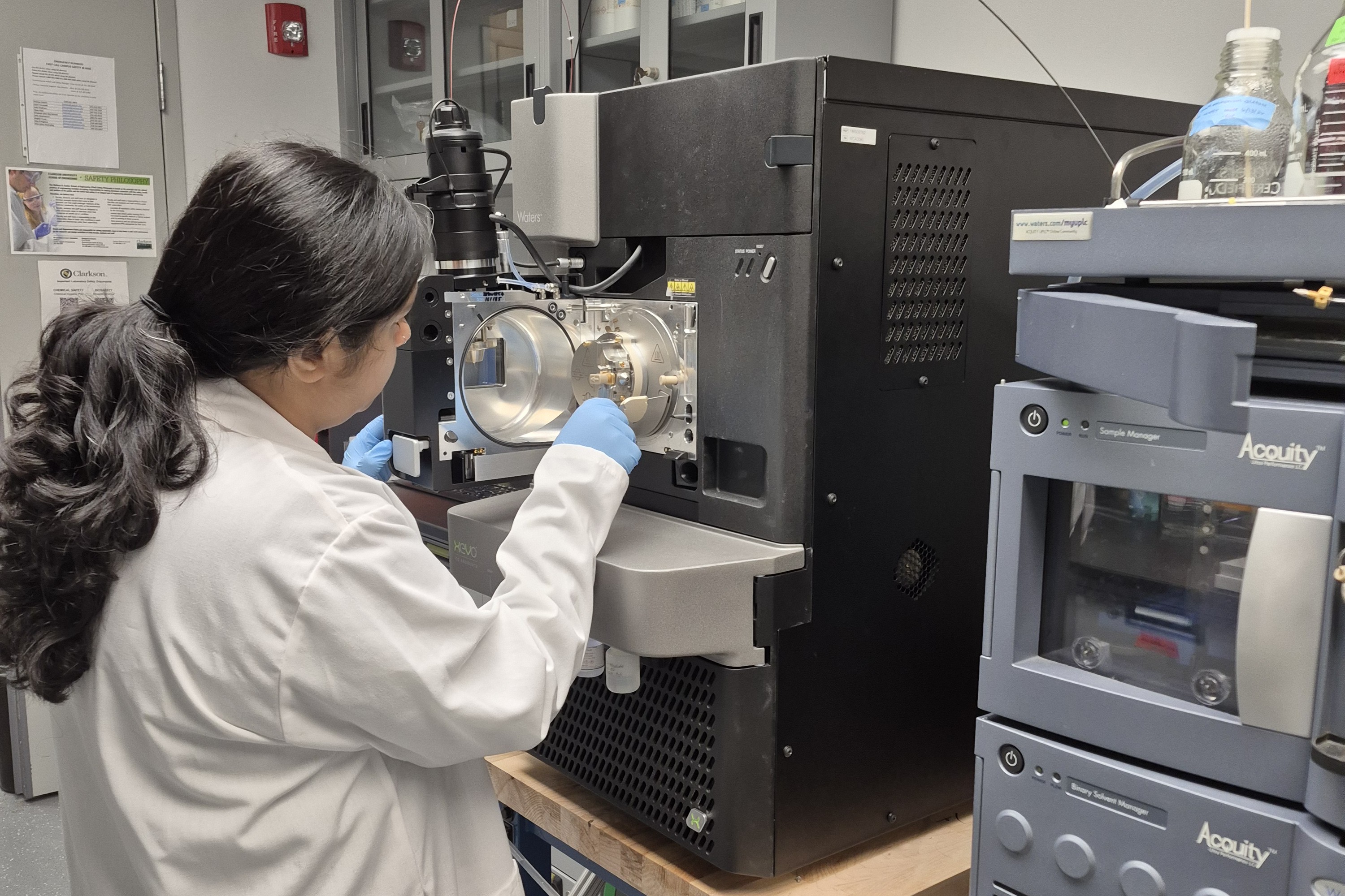 A Clarkson University researcher in a white lab coat and blue protective gloves calibrating a mass spectrometer. A Waters ACQUITY UPLC system is visible stacked in the foreground on the right side of the laboratory bench.