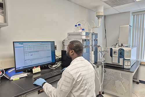 A researcher wearing a white lab coat and blue gloves sits at a computer workstation, analyzing data next to a liquid chromatography mass spectrometry setup in a laboratory.