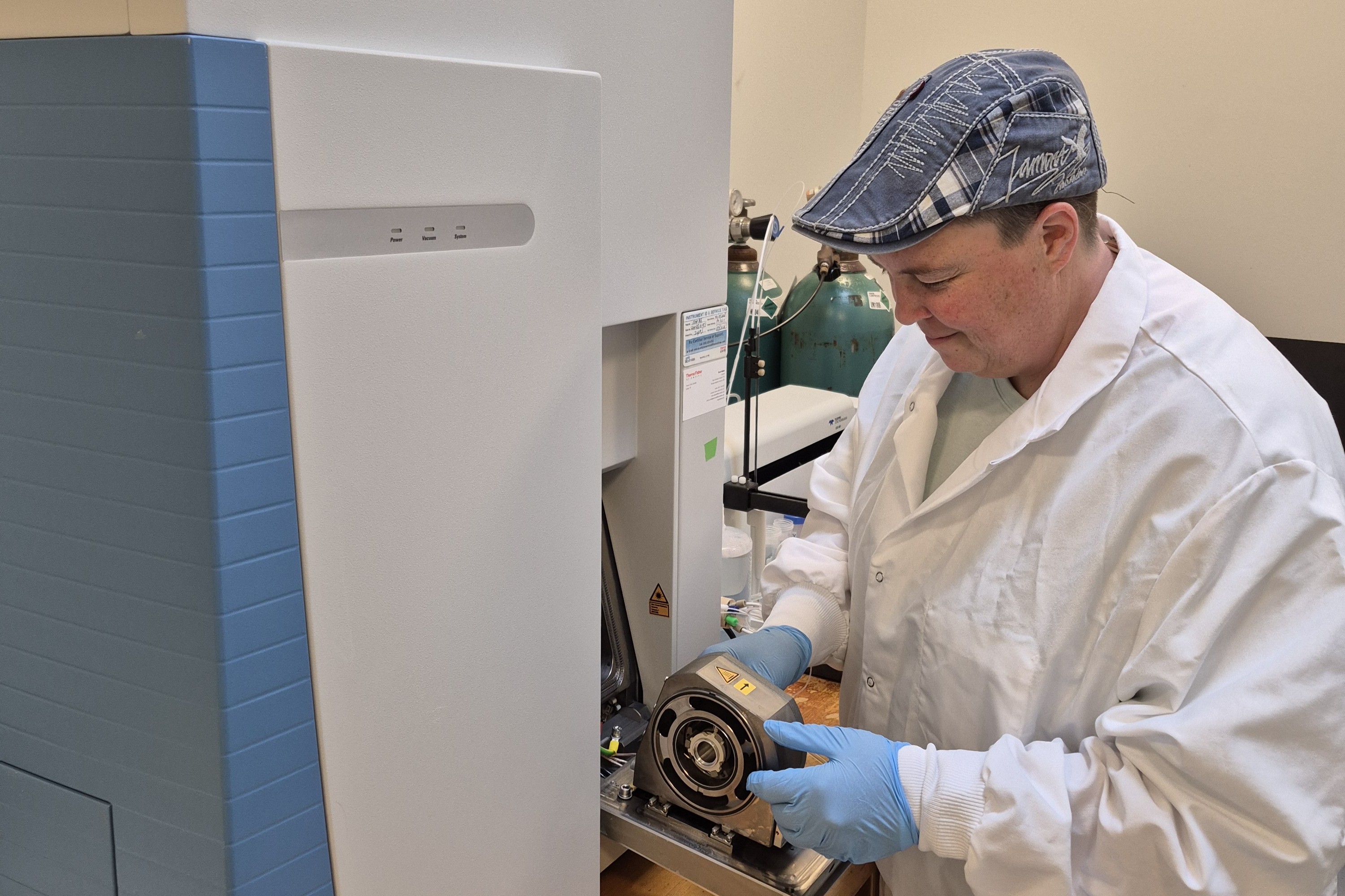 A Clarkson University researcher wearing a white lab coat, blue gloves, and a patterned flat cap adjusting a circular component on a large laboratory analytical instrument. Gas cylinders are visible against the wall in the background.