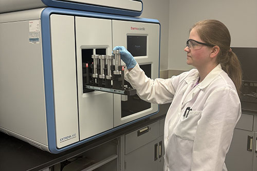 A researcher wearing a white lab coat, safety glasses, and blue gloves adjusts sample cylinders on an automated tray connected to a large analytical instrument in a laboratory.