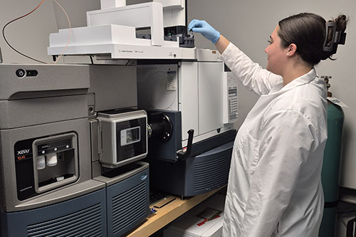 A researcher wearing a white lab coat and blue gloves places a sample vial into the autosampler tray of a high-performance liquid chromatography mass spectrometer in a lab.