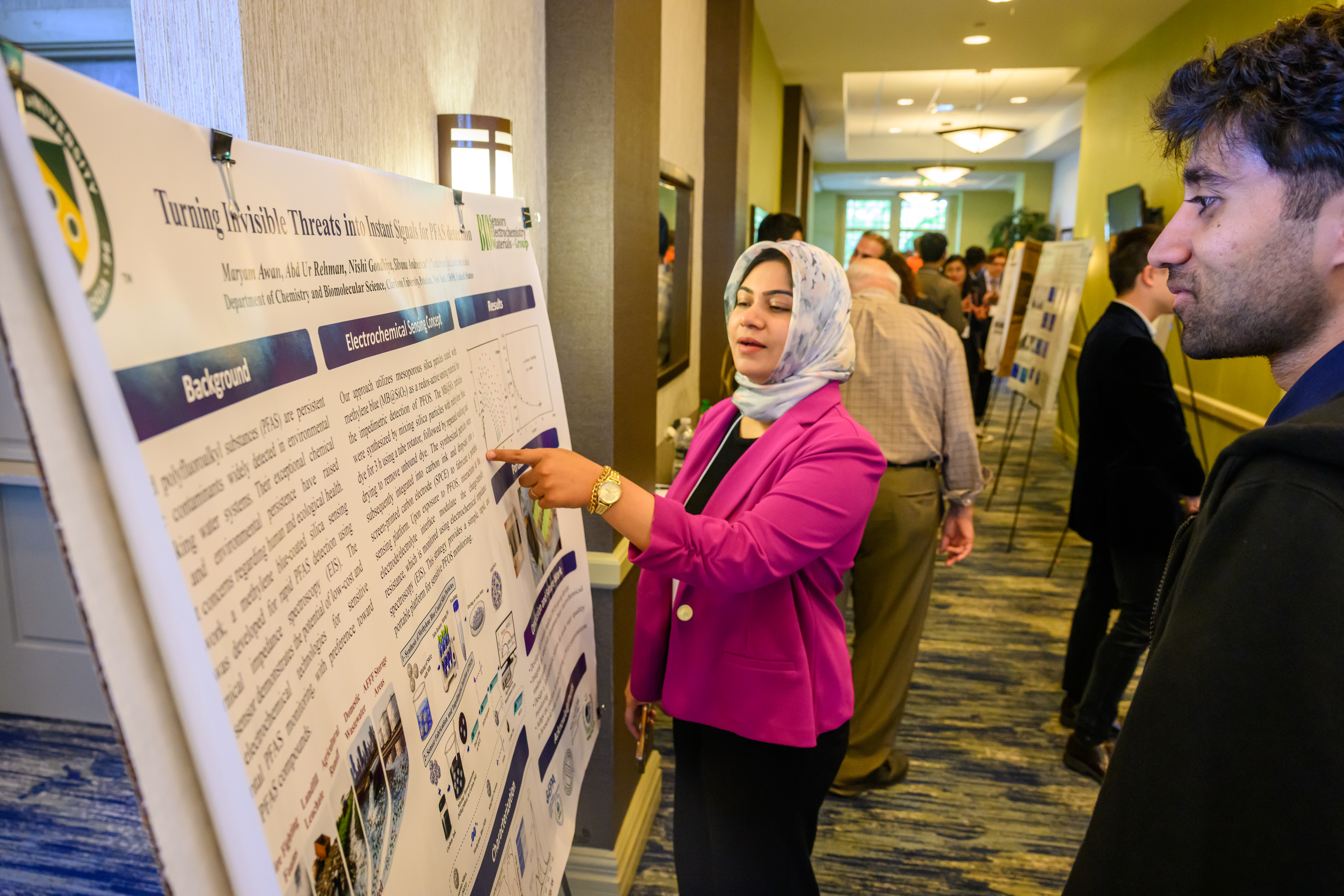 A person wearing a bright pink blazer and patterned headscarf presents a research poster at an academic conference, pointing to information on the display while speaking with another attendee. The poster contains scientific text, charts, and diagrams related to environmental sensing research. Several other participants and poster presentations are visible in the background along a hotel or conference center hallway, creating a busy research showcase environment.