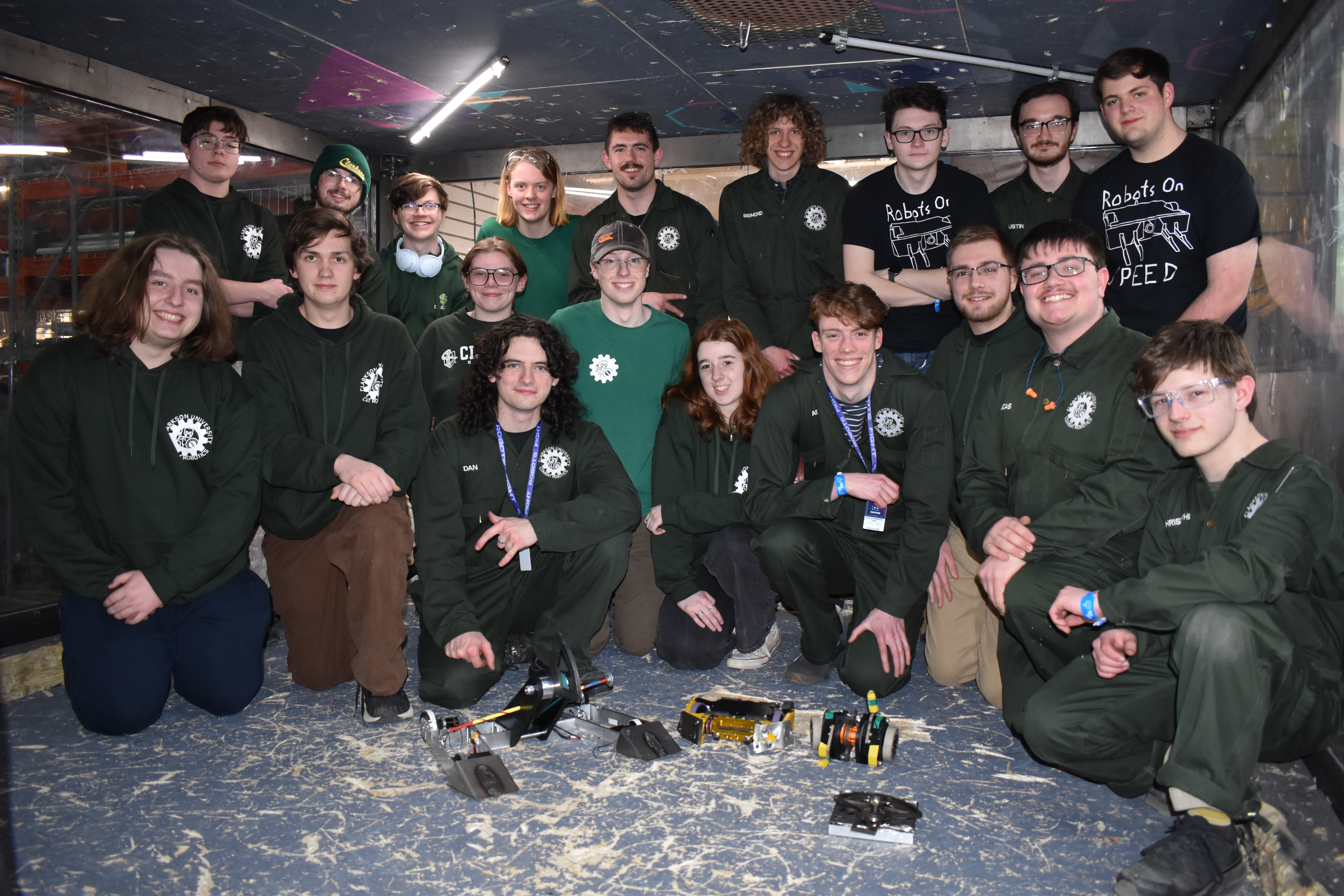 group of about 15 Clarkson University Combat Robotics Team students wearing green team apparel, posing together inside an enclosed combat arena. Several small, custom-built combat robots rest on the wood-shaving-covered floor in front of them.