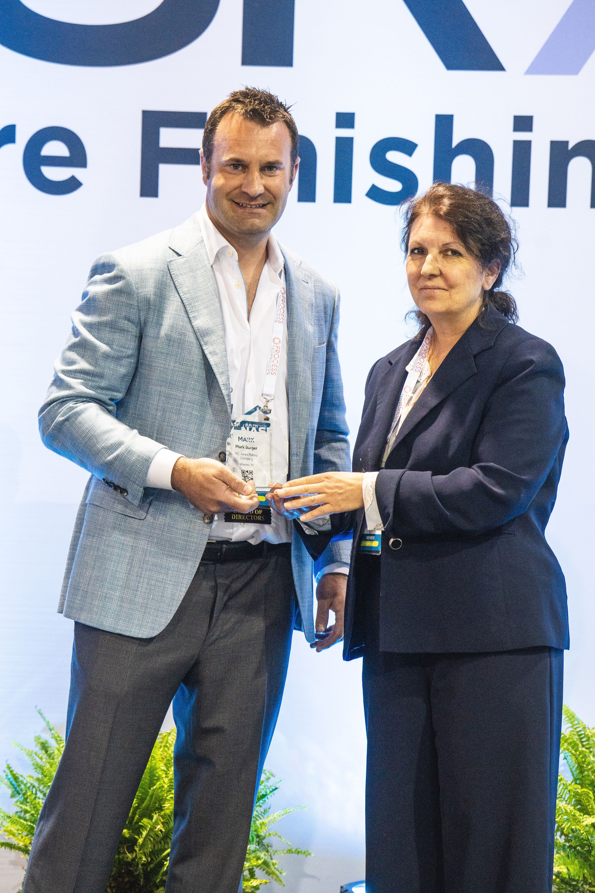 Two people stand on a stage during a professional conference or awards ceremony, jointly holding a plaque or award. One person wears a light blue blazer and conference badge, while the other wears a dark business suit. A large event backdrop with partially visible text and conference branding is behind them, with decorative plants along the front of the stage. Both are facing the camera during the award presentation.