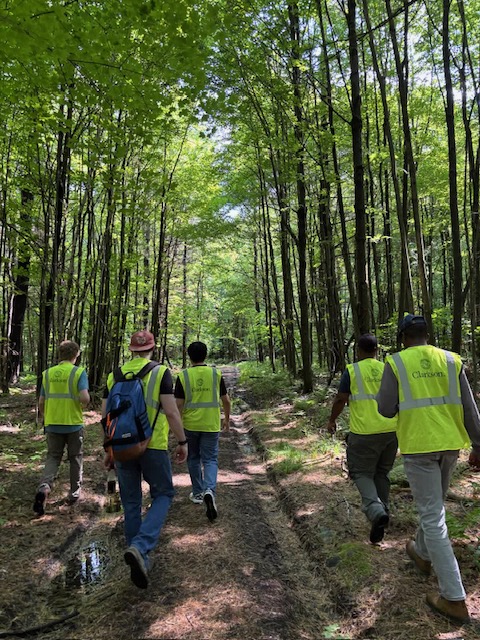Five people wearing high-visibility safety vests walk along a wooded trail through a dense forest. Sunlight filters through the green tree canopy overhead, and the group follows a narrow path with muddy tire tracks and patches of standing water. Some of the vests display the Clarkson University logo.