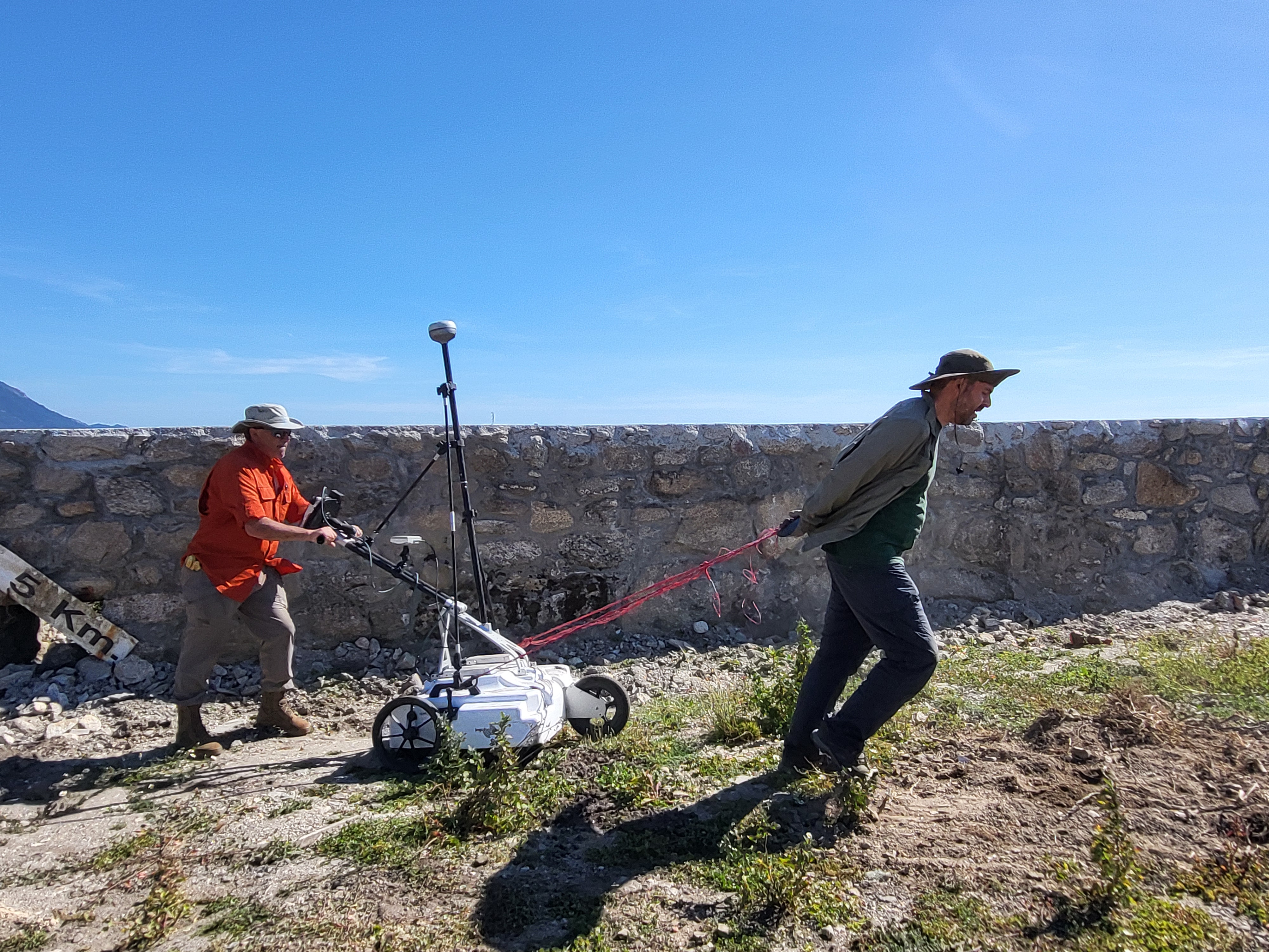 Two researchers conduct a ground-penetrating radar survey at an outdoor archaeological site. One researcher pulls a wheeled radar unit with ropes while the other guides and monitors the equipment from behind. A stone wall runs along the background under a clear blue sky, highlighting the use of non-invasive geophysical technology to investigate buried archaeological features.