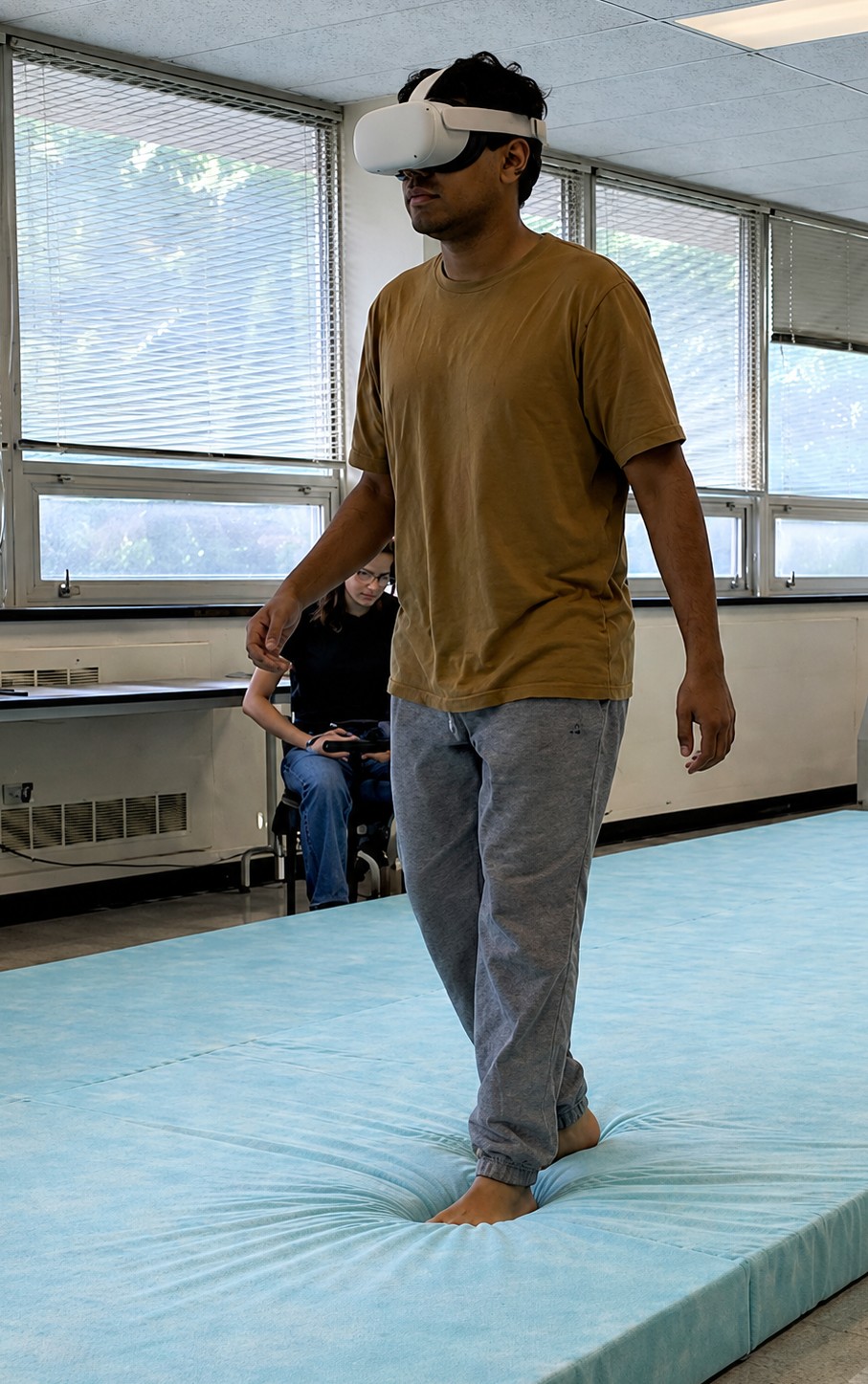 A person wearing a virtual reality headset walks through a laboratory during a gait assessment. Large windows line the room, and another person observes from the background. The VR headset is used as part of a walking balance evaluation that measures how visual information affects balance during movement.
