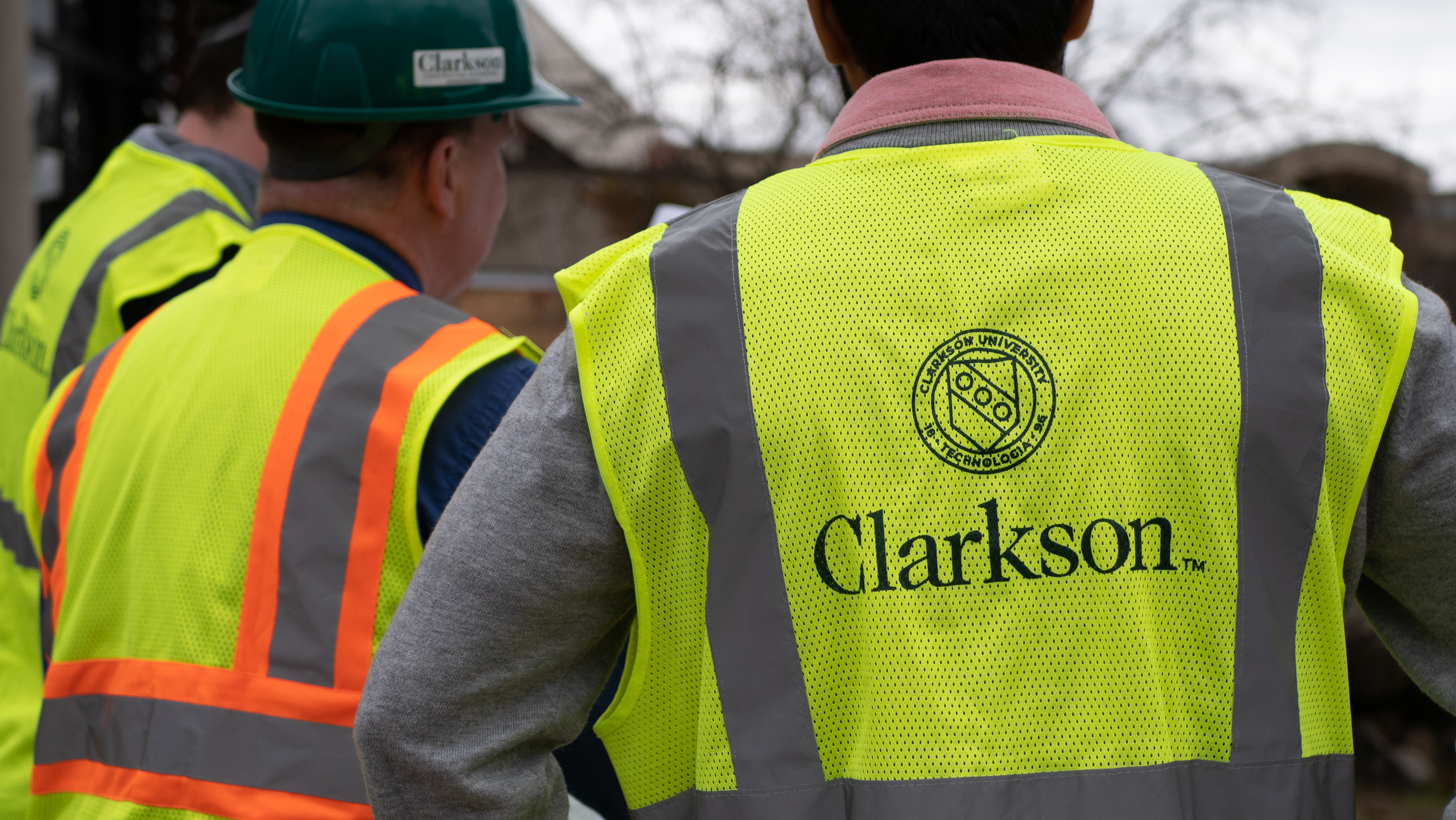Three people wearing high-visibility safety vests at a work site, including one with the Clarkson University logo and another wearing a green Clarkson hard hat.