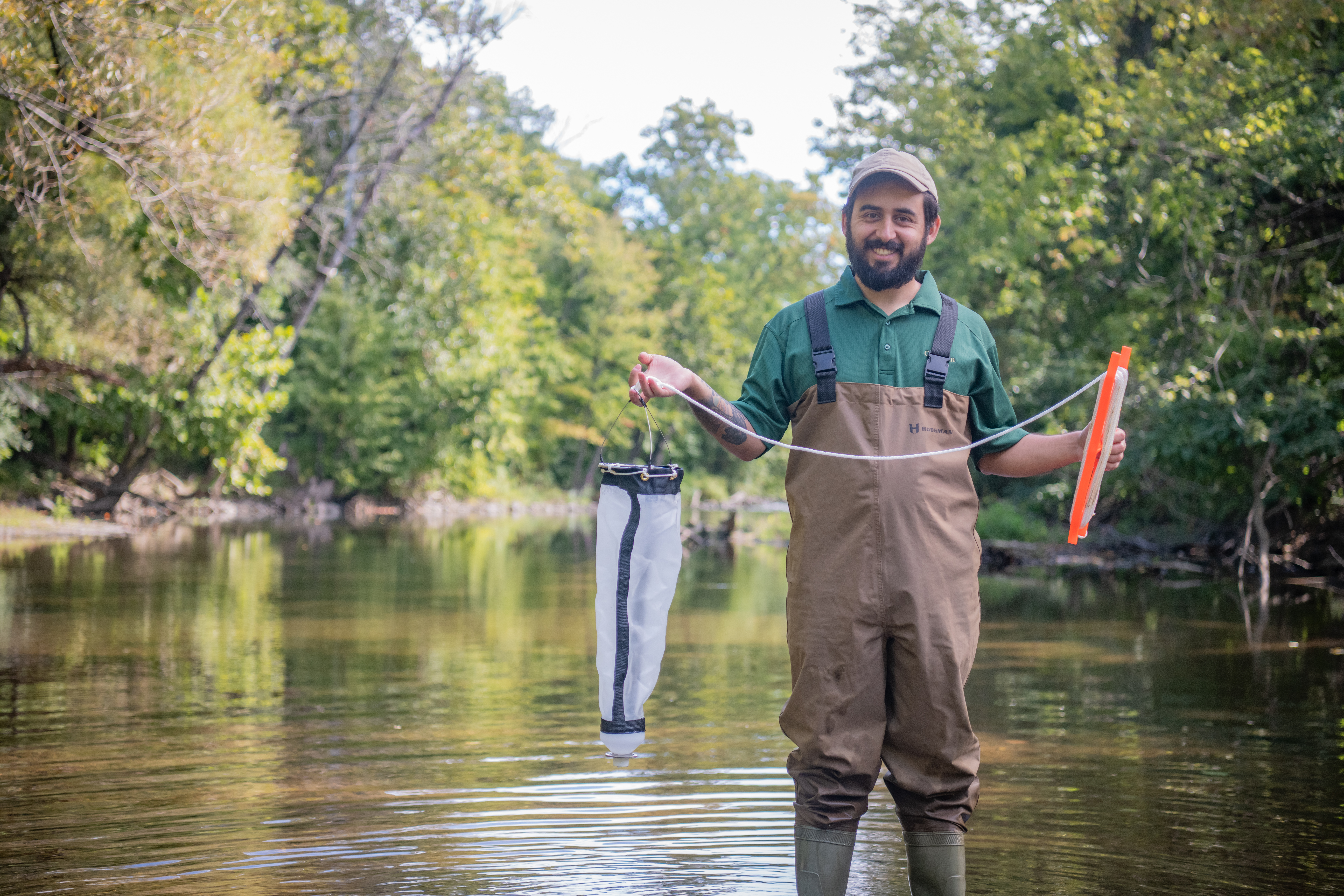A researcher wearing chest waders stands in a shallow, tree-lined stream holding water-sampling equipment used for environmental research.