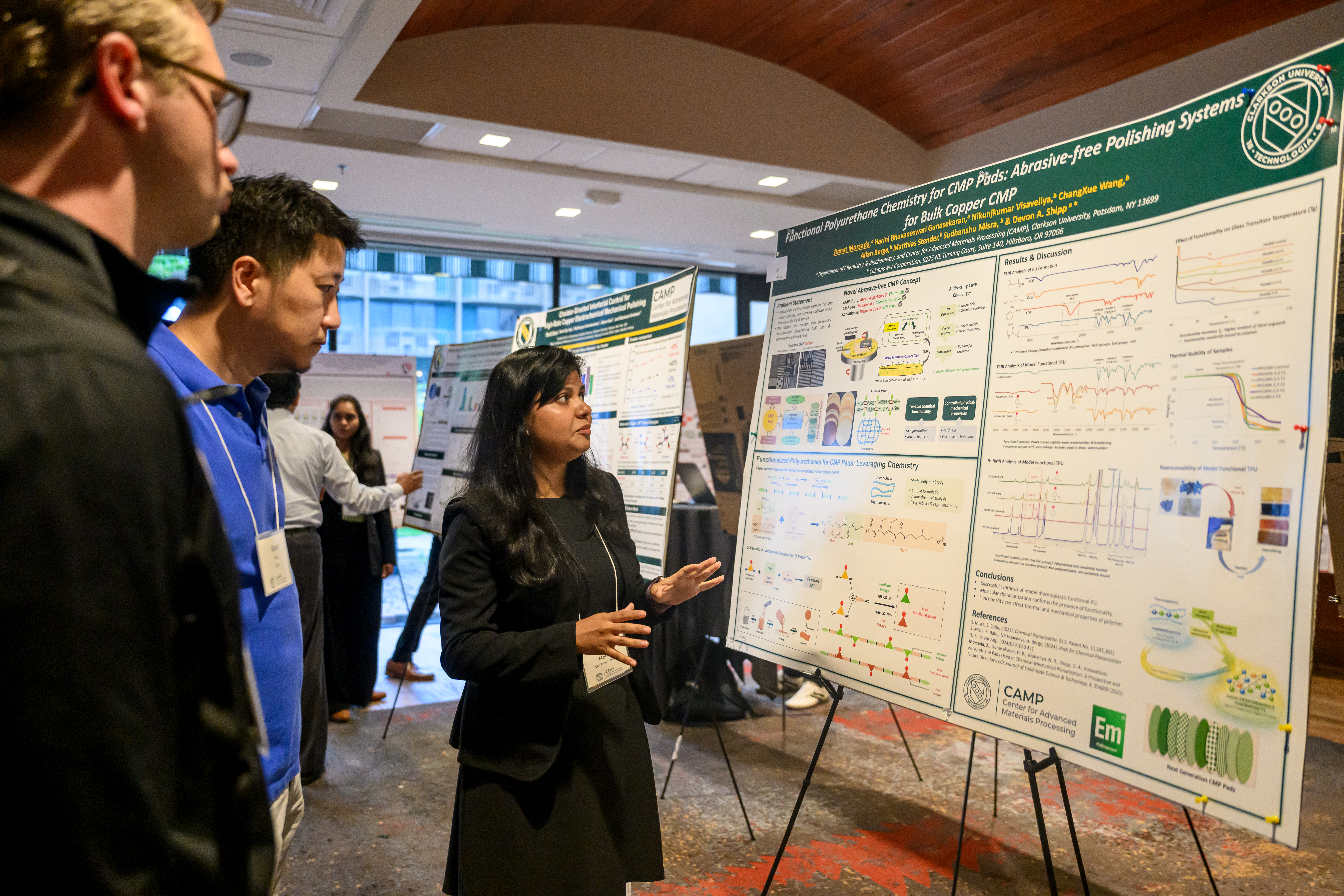 A researcher presents a scientific poster on chemical mechanical planarization to attendees at Clarkson University’s CAMP CMP Symposium, with additional research posters displayed in the background.