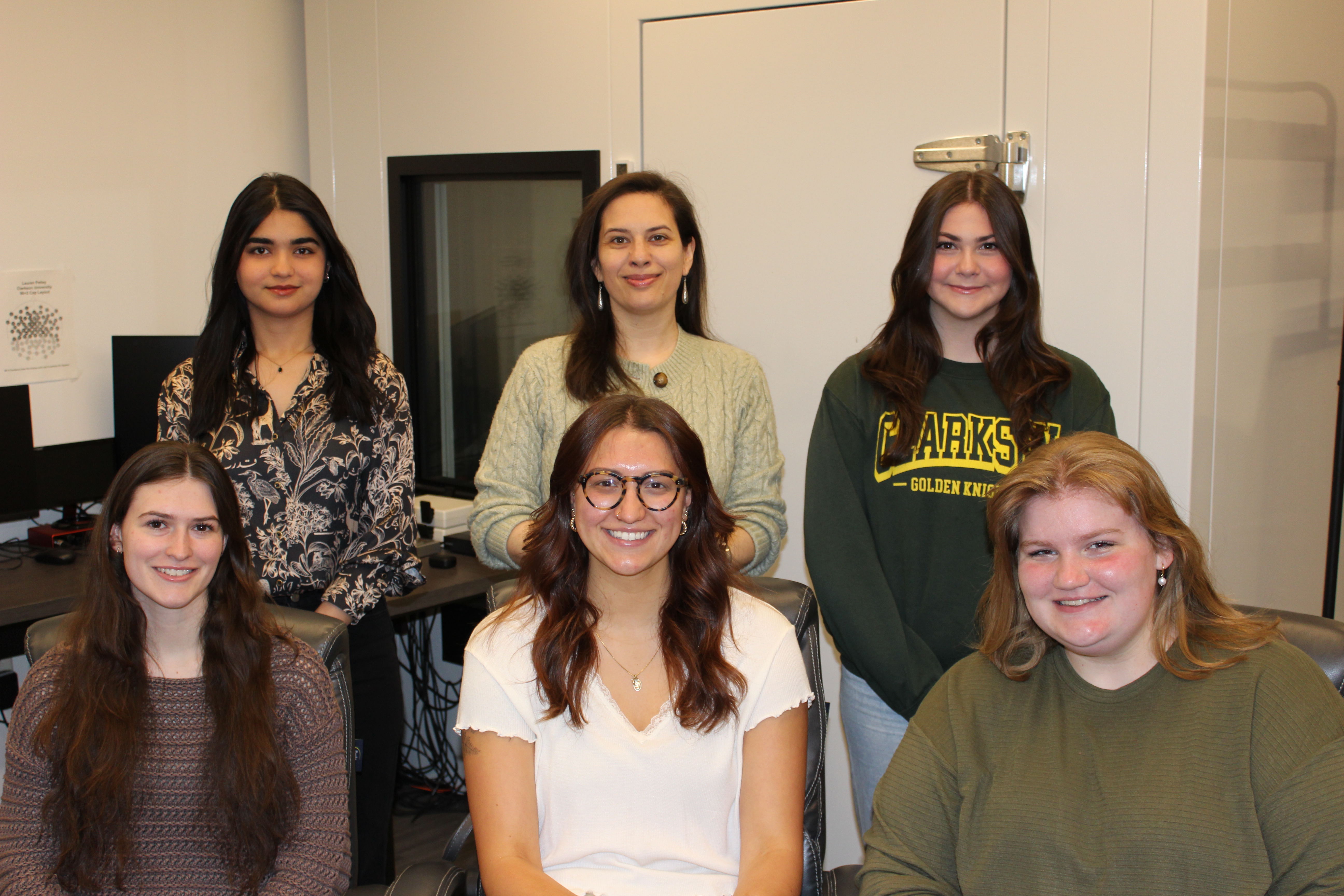 Six female students and faculty gathered indoors for a group portrait, with three seated in front and three standing behind them.