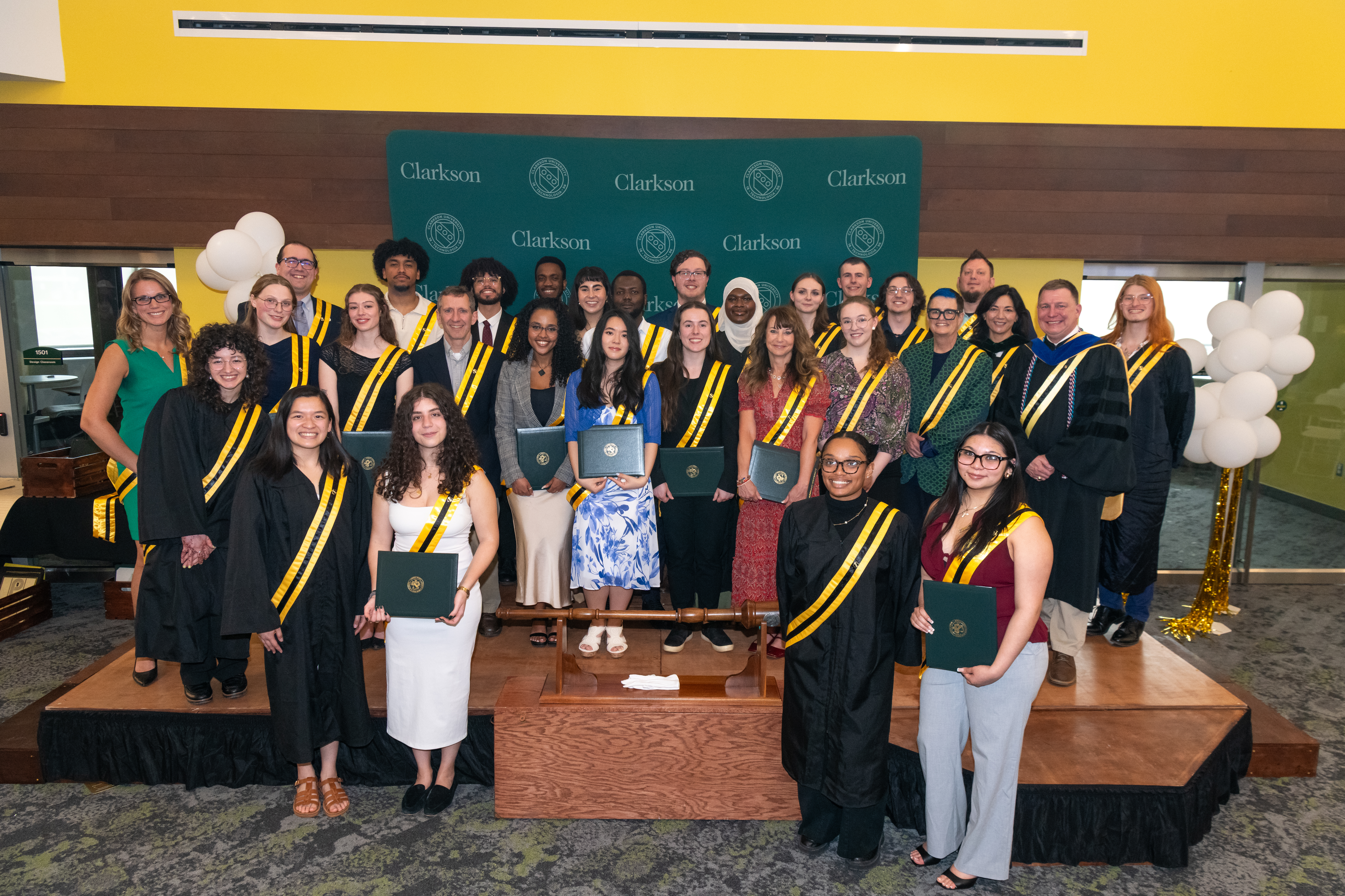 A group photo of students and faculty on a wooden stage at a Clarkson University ceremony, wearing black graduation gowns or formal attire with green and gold striped sashes while holding dark green certificate folders in front of a green backdrop.