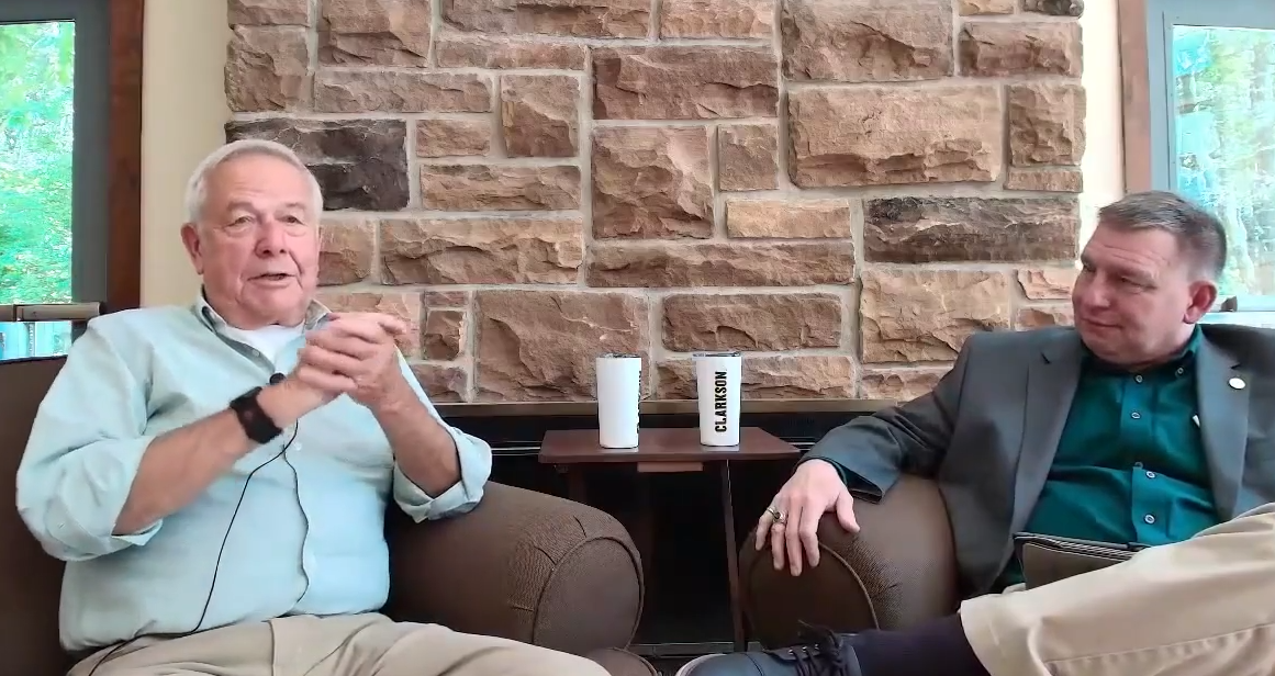 Two men sit in armchairs and talk in front of a stone wall during an interview-style conversation, with Clarkson-branded drinkware on a table between them.